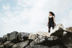 A woman stands defiant on large boulders. Her pants are in tatters, yet she's a survivor.